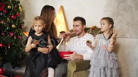 幸福的四口之家坐在圣诞树旁吃巧克力饼干可爱的母亲父亲和两个女儿视频的预览图