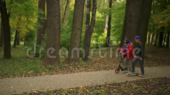 两个青少年骑滑板车和一个便士板他们沿着公园的小路前进公园里的秋日绿色视频的预览图
