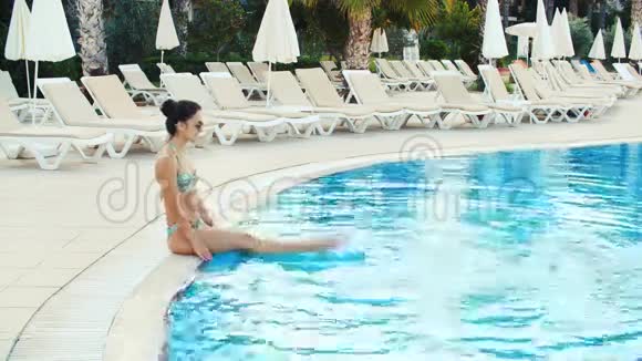 年轻的黑发女人穿着比基尼戴着太阳镜坐在游泳池里在阳光明媚的夏日泼水视频的预览图
