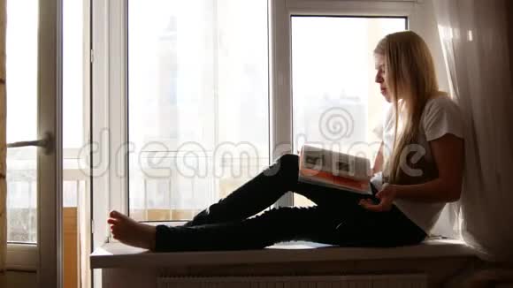 坐在窗台上翻书的漂亮金发女孩视频的预览图