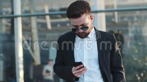 一个戴太阳镜的年轻胡子男子站在机场候机楼旁用他的手机旋转商务视频的预览图