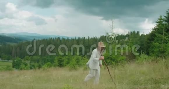 穿着旧衣服的老灰人拿着拐杖走路青山的壮丽景色视频的预览图