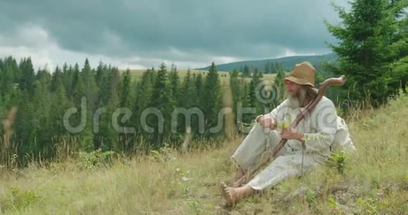 穿着旧衣服的老人坐在山上的草地上闻着花横向视野视频的预览图