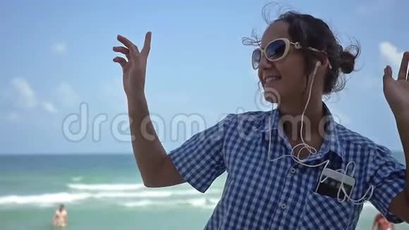 年轻漂亮的女士戴着太阳镜跳舞一边在海边的海滩上用手机听音乐慢动作1920x1080视频的预览图