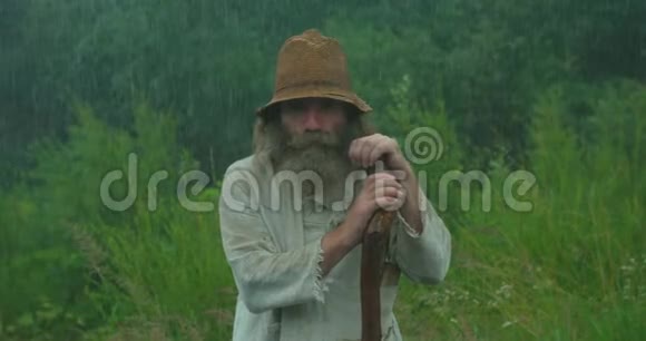这位穿着旧衣服和旧帽子的老人在雨中靠在拐杖上咀嚼视频的预览图