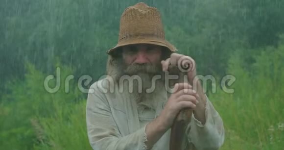 有着长长的灰色胡须和旧衣服的讨人喜欢的老人在雨中靠在拐杖上咀嚼着特写视频的预览图