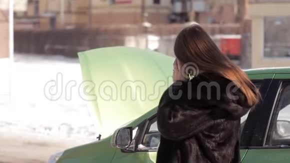 冬天一个怀孕的女孩站在一辆有故障的车旁边车上开着一个引擎盖试图用手机给拖车打电话视频的预览图