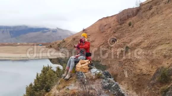 一个女孩坐在湖边的一块岩石上的鸟瞰图在电话里拍下了一张风景照片并拍摄了一张照片视频的预览图