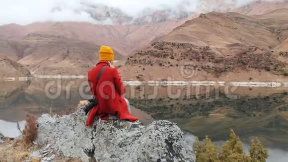 一个女孩坐在湖边的一块岩石上的鸟瞰图在电话里拍下了一张风景照片并拍摄了一张照片视频的预览图