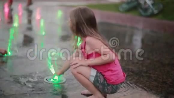 在炎热的夏天可爱的小女孩蹲在喷泉边玩彩色喷水视频的预览图