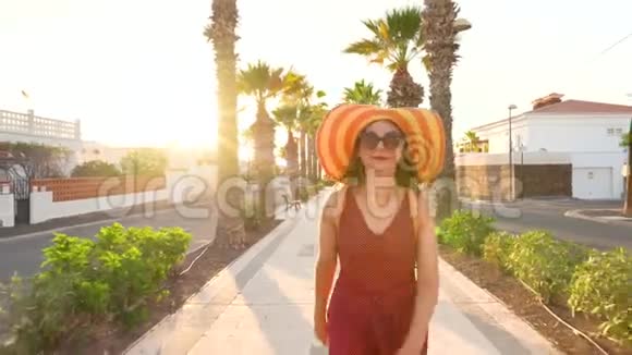 日落时分戴着一顶黄色大帽子的幸福女人在棕榈巷散步视频的预览图