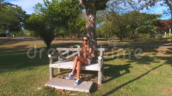 空中女孩坐在公园的长凳上用智能手机打字慢镜头飞走高清镜头视频的预览图