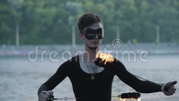 身着黑色衣服戴着面具技艺精湛身材苗条的年轻男子的肖像在河岸上用火焰表演视频的预览图