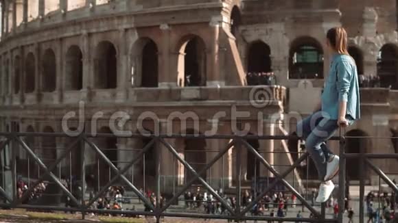 女人坐在栅栏上看着意大利罗马的斗兽场摆动着她的腿人群靠近视线慢动作视频的预览图