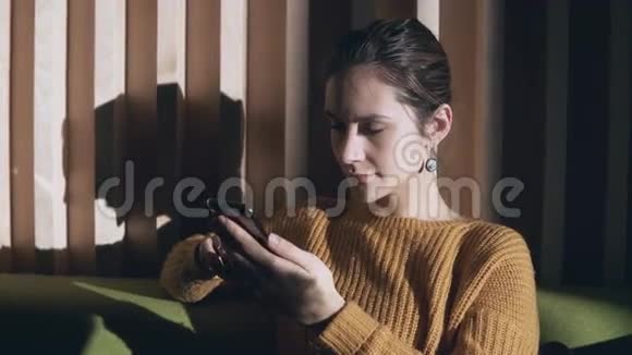 年轻女子坐在客厅的沙发上使用智能手机严肃的女性早上浏览互联网视频的预览图