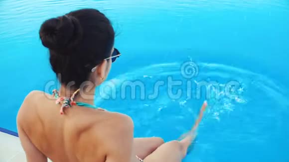 年轻的黑发女人穿着比基尼戴着太阳镜坐在游泳池里在阳光明媚的夏日泼水视频的预览图