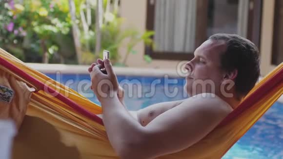 光着胸的胖子躺在吊床上在游泳池旁用手机慢动作度假时放松一下视频的预览图