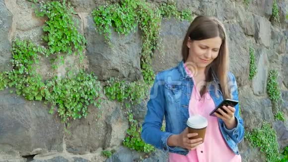 穿着棉袄用智能手机喝咖啡的漂亮女孩背景中野石的复古墙视频的预览图