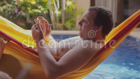 胖懒人光着胸膛躺在吊床上用手机在游泳池边看书3840x2160视频的预览图