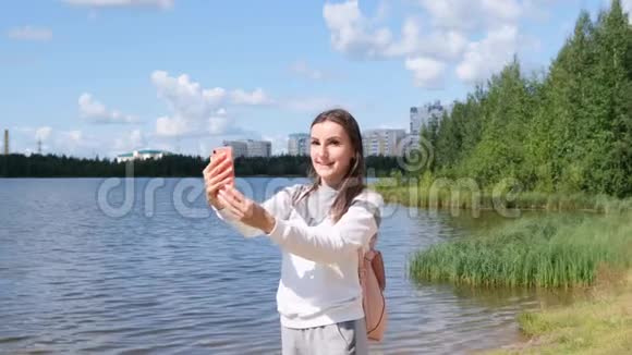 带背包的漂亮女孩在湖边自拍并在Skype上用手机交谈视频的预览图
