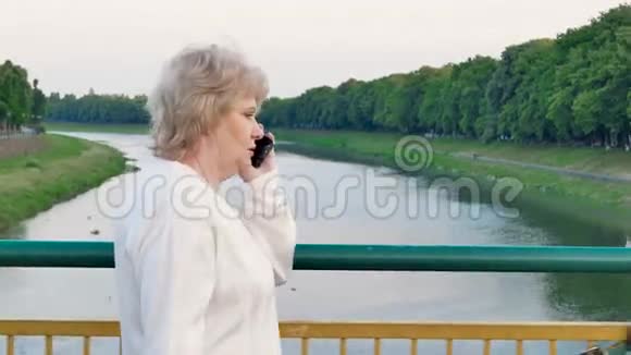 一位年长的女士穿过河上的桥在智能手机上交谈视频的预览图