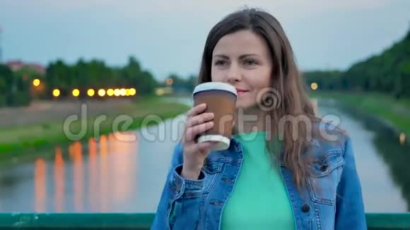 穿着棉袄站在河边的桥上喝咖啡的漂亮姑娘视频的预览图