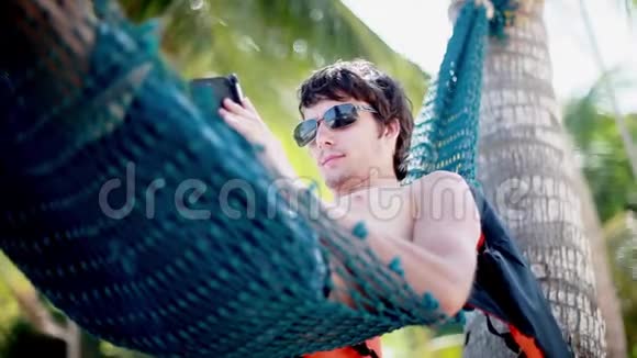戴太阳镜的年轻人躺在吊床上在美丽的异国海滩上使用智能手机1920x1080视频的预览图