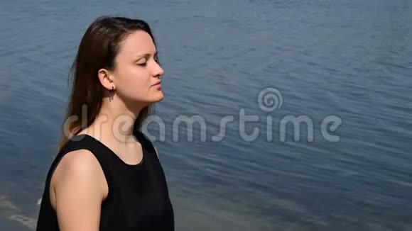 美丽的黑发女人闭着眼睛穿着一件黑色衬衫在海岸边享受视频的预览图