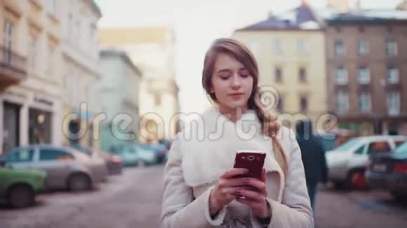 年轻漂亮的金发女郎在她的手机上打字而走在寂寞的街道上在阳光明媚的早晨环顾四周视频的预览图