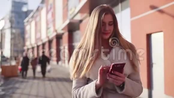 年轻漂亮的金发女郎走在市中心一座现代化建筑附近的繁华街道上一边发短信视频的预览图