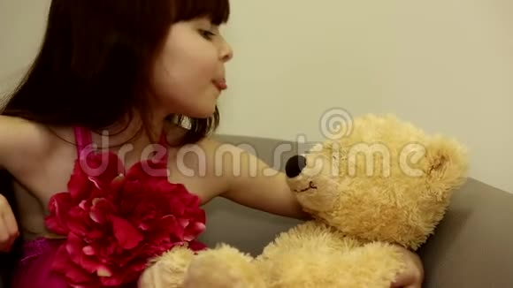 快乐可爱的小女孩安静的小孩玩着露出舌头的样子亲吻她在椅子上的毛绒玩具熊视频的预览图