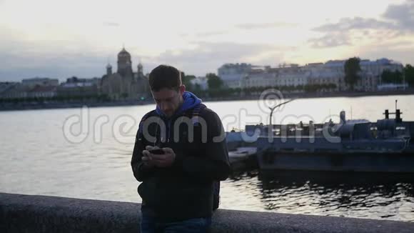 一个年轻人拿着电话站在河堤上在日落的背景下写着一条信息视频的预览图