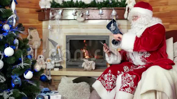 圣诞老人使用平板电脑房间内有壁炉圣尼古拉斯坐在椅子上有电子设备视频的预览图