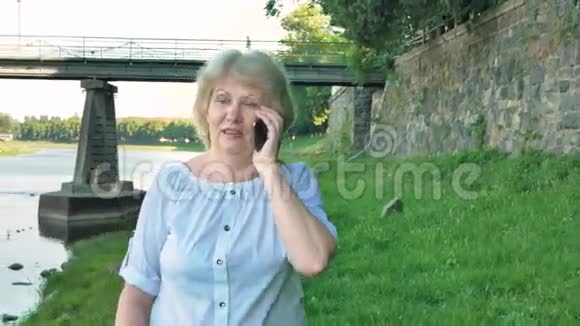 老年妇女沿着海滨散步在智能手机上交谈背景中的野石桥和老墙视频的预览图