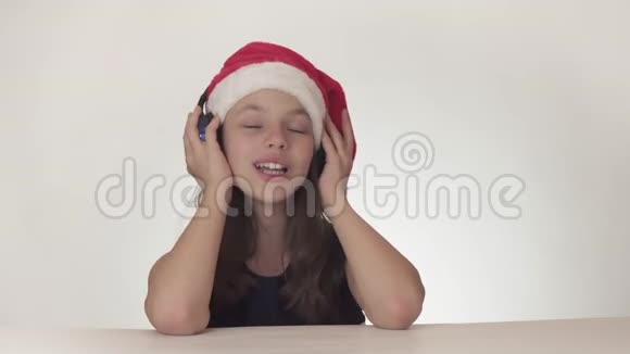 戴着圣诞老人帽子的美丽顽皮的少女在耳机上听音乐在白色背景上唱歌视频的预览图