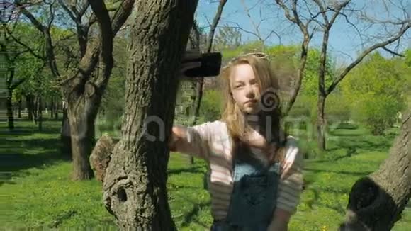 一个十几岁的女孩坐在一棵树上拿着一部电话一个穿破牛仔裤的女孩自拍公园里漂亮的女孩视频的预览图