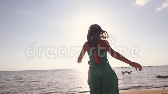 穿着绿色衣服在海滩上奔跑的漂亮年轻女子慢悠悠地旋转着举起双臂享受自由视频的预览图