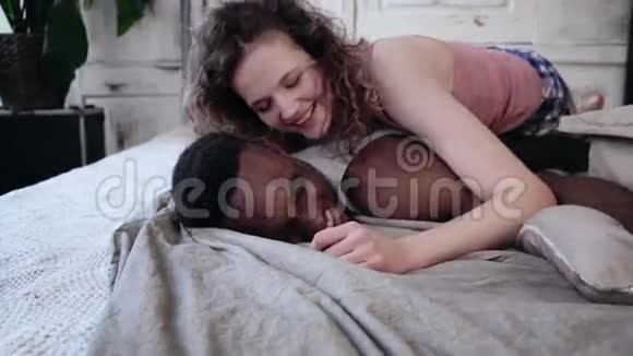 年轻漂亮的女人躺在穿睡衣的男人身上多种族夫妇抱在床上手牵着手微笑着视频的预览图