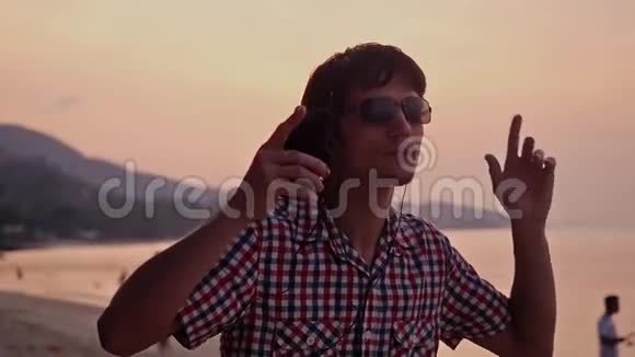 年轻英俊的快乐男子戴着太阳镜听音乐用手机在热带海滩跳舞在惊人的时候视频的预览图