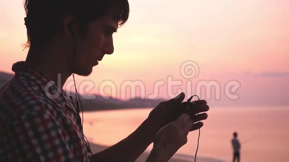 年轻的帅哥拿着手机在热带海滩听音乐在惊人的日落慢动作1920x1080视频的预览图