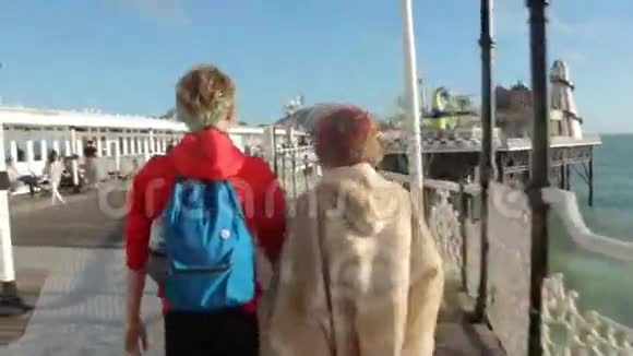 英国布莱顿漂亮的年轻夫妇正沿着码头散步年轻人牵手在海洋海岸视频的预览图