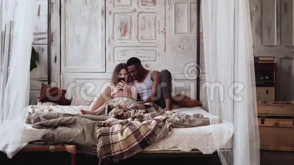 床上穿睡衣的多种族夫妇使用智能手机非洲男人和欧洲女人浏览互联网视频的预览图