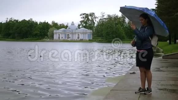 女孩站在湖边公园的雨伞下在电话里拍照慢动作1920x1080满的视频的预览图