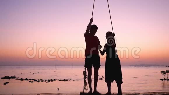 这对年轻夫妇站在一个秋千上手牵着手在热带海滩上对抗大海在令人叹为观止的日落慢动作视频的预览图