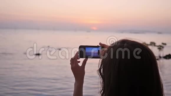 女人在海边海滩上美丽的日落度假时用智能手机拍照慢动作视频的预览图