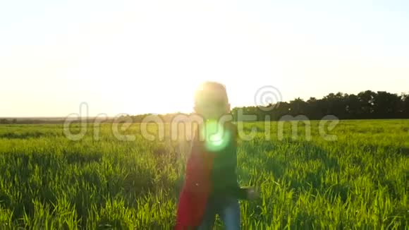 日落时一个穿着超级英雄服装穿着红色雨衣的快乐孩子穿过绿草摄像机移动视频的预览图