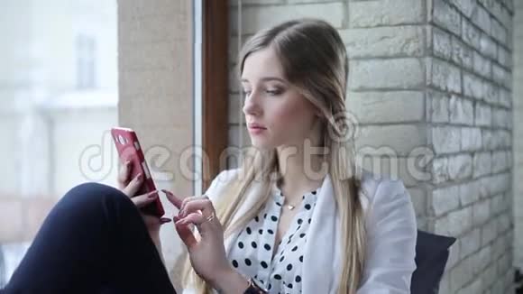 漂亮的年轻商务女性坐在靠近窗户的灯光办公室里在手机上打字视频的预览图