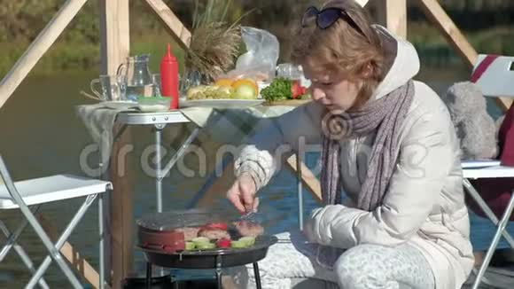 年轻女子穿着温暖的衣服在烤架上准备蔬菜和肉一只狗在附近散步在河岸上野餐视频的预览图