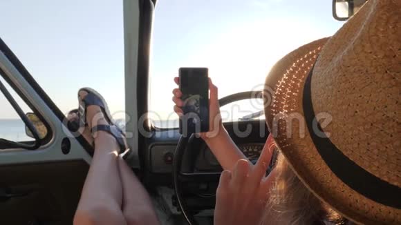 美丽的女孩在背景海的老式汽车上在手机上自拍快乐的女性在汽车上做自拍视频的预览图