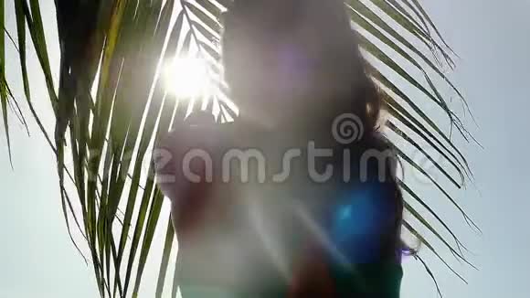 女孩穿过一棵棕榈树的树枝来到海边阳光透过棕榈树照射视频的预览图